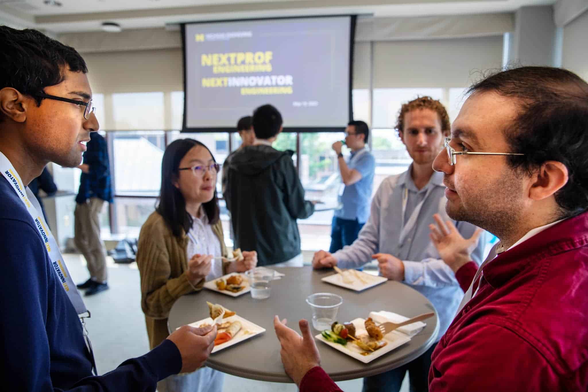 Group of four people chatting around a standing table while eating. Projection screen in background reads "NextProf Engineering" and "NextInnovator"