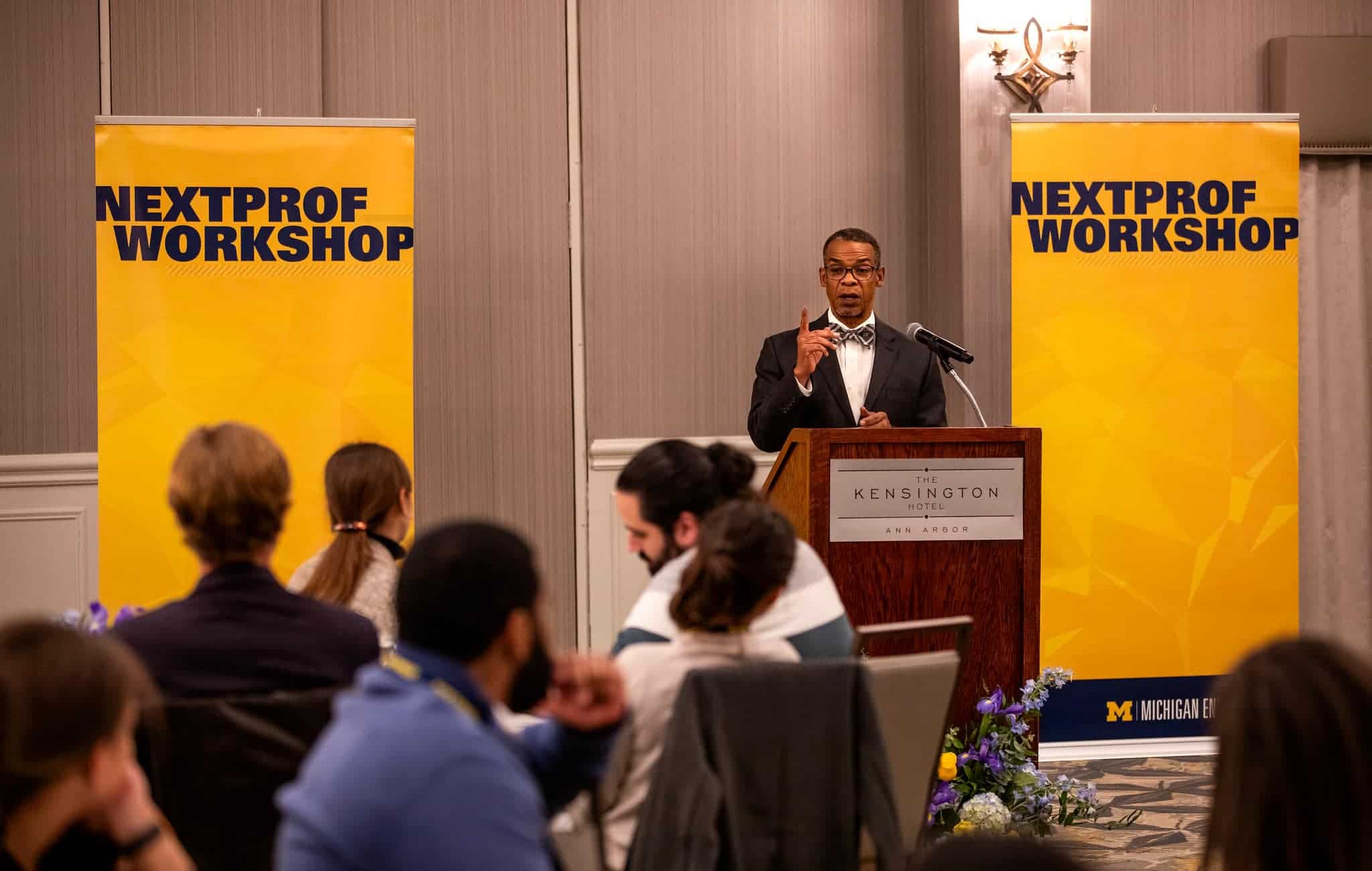 Black man at podium between two bright yellow banners with the words "Nextprof workshop" on both